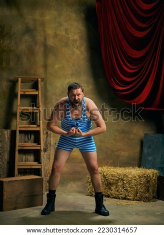 Winner emotions. Mature man, retro circus strongman wearing striped sports swimsuit shouting isolated over dark vintage circus backstage background. Art, 30s, 40s fashion style and inspiration