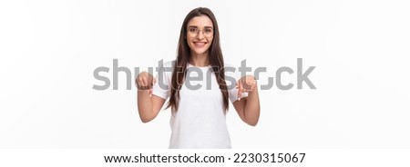 Click here. Portrait of brunette gorgeous young woman in glasses, pointing fingers down to show advertisement, smiling pleased, suggest visit link, recommend download or subscribe, white background. Royalty-Free Stock Photo #2230315067