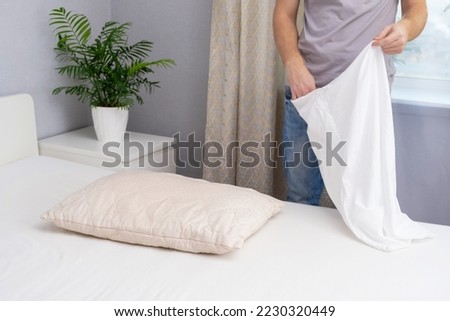A man tucks a pillow with a fresh bright white pillowcase. Making the bed with fresh bed linen by a white man. The day of the change of bed linen. The day of washing bed linen in the laundry room. Royalty-Free Stock Photo #2230320449