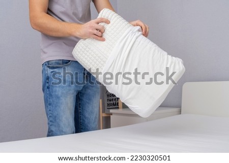 A man tucks an orthopedic pillow with a fresh bright white pillowcase. Making bed with fresh bed linen by white man. The day of change of bed linen. The day of washing bed linen in the laundry room. Royalty-Free Stock Photo #2230320501