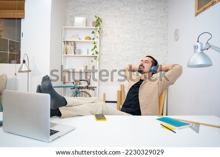 Positive young man keeping hands behind head while listening to music in headphones at home. resting after work in his studio. Creative designer thinking in new ideas. High quality photo