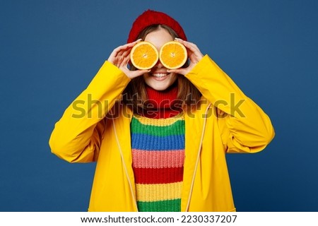 Young happy fun woman wear sweater red hat yellow waterproof raincoat outerwear cover eyes with orange halves isolated on plain dark royal navy blue background Outdoors wet fall weather season concept