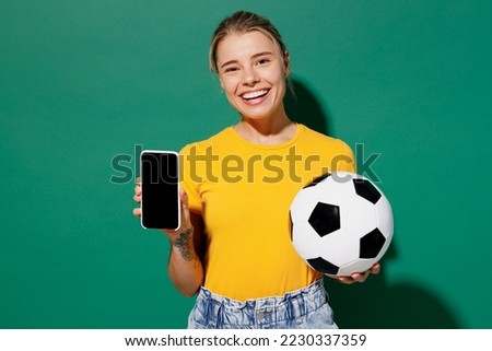 Young happy woman fan wear basic yellow t-shirt cheer up support football sport team hold soccer ball watch tv live stream use mobile cell phone blank screen isolated on dark green background studio