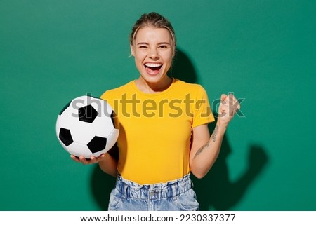Young woman fan wear basic yellow t-shirt cheer up support football sport team hold in hand soccer ball watch tv live stream do winner gesture celebrate clench fist isolated on dark green background Royalty-Free Stock Photo #2230337377