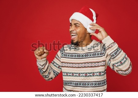 Side view merry young man wear cozy Christmas sweater Santa hat headphones listen music look aside clench fist posing isolated on plain red background. Happy New Year 2023 celebration holiday concept