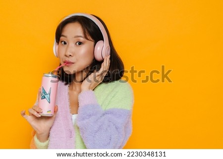 Young cute asian woman in headphones drinking soda isolated over yellow studio background Royalty-Free Stock Photo #2230348131