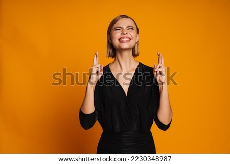 Young beautiful woman with closed eyes doing hope gesture with crossed fingers both raised hands standing over isolated orange studio background