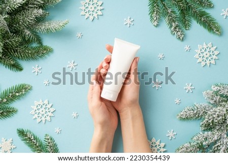 Winter season skin care concept. First person top view photo of young woman's hands holding white tube without label over snowflakes and pine branches in frost on isolated pastel blue background Royalty-Free Stock Photo #2230354439