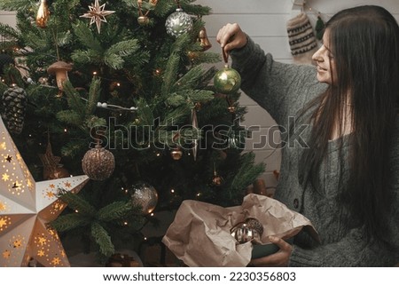 Stylish woman in cozy sweater decorating christmas tree with stylish bauble in atmospheric festive room. Merry Christmas! Winter holidays preparation. Decorating xmas tree with vintage toy