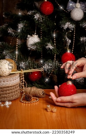 Female hands making Christmas decorations on the table next to year tree as a background, vertical photo, selective focus. Handmade balls, New Year, holiday, atmospheric, craft, winter concept