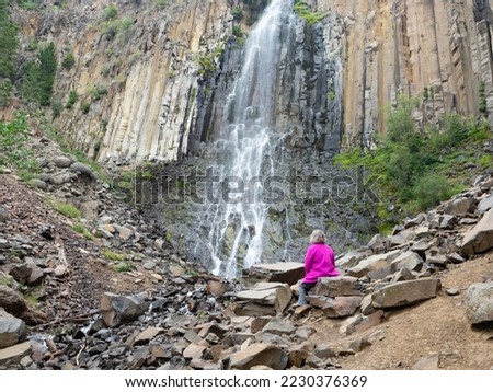 Senior adult woman wearing a fuchsia jacket and gray pants sitting on a boulder at the base of Palisade Falls in Hyalite Canyon, Montana, on an overcast day. Royalty-Free Stock Photo #2230376369