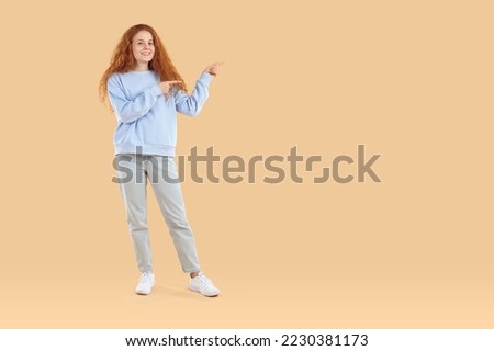 Full length portrait of positive redhead girl in blue sweatshirt and jeans is pointing index fingers on copyspace on beige background. Banner for advertisement, sales, marketing, good offer concept.