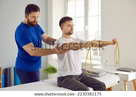 Physiotherapy specialist and his male patient use special equipment during rehabilitation. Young man doing remedial physical exercises with resistance band under supervision of serious physiotherapist Royalty-Free Stock Photo #2230381175
