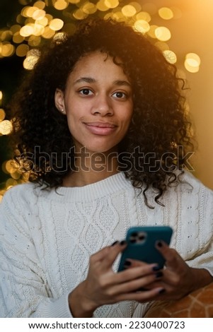 Merry Christmas and Happy Holidays! Young african american woman chatting on a smartphone on by the Christmas tree in the cozy living room of the house                              