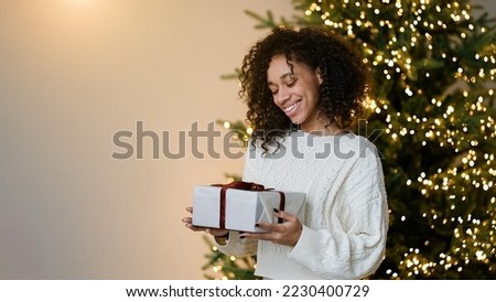 Merry Christmas and Happy Holidays! Young african american woman holding a holiday gift on by the Christmas tree in the cozy living room of the house, the concept of happiness                         