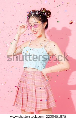 Portrait of beautiful young Asian happy woman in blue cute fashion dress wear sunglass hands raised celebrate confetti fly fall on isolated pastel pink color background empty space. Party concept.