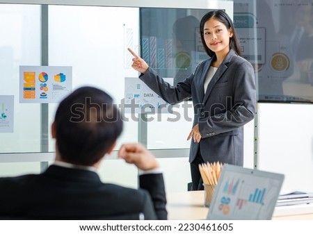 Millennial Asian successful professional female businesswoman lecturer in formal suit standing smiling pointing finger at graph chart data information on glass board presenting explaining to audience. Royalty-Free Stock Photo #2230461605