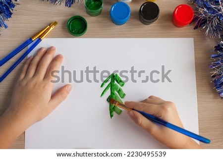 Children's hands of a girl dip a brush in paint. In the background there are brushes, paints and New Year's tinsel. Paints on the table. A child draws and makes crafts in kindergarten
