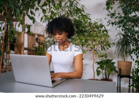 Serious curly African-American young woman working typing on laptop computer sitting at desk in home office room with modern biophilia design, on background of green plants. Concept of house garden. Royalty-Free Stock Photo #2230499629