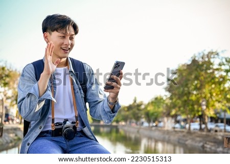 Handsome and happy young Asian male traveler smiling, waving hand, video call with his friend through smartphone while enjoys strolling around the city.