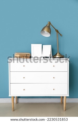 Blank frames, lamp and books on chest of drawers near blue wall