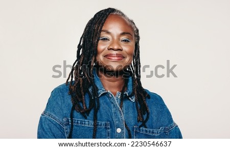 Portrait of a woman with dreadlocks looking at the camera with a smile while standing against a grey background. Stylish mature woman wearing a denim jacket and make-up in a studio.