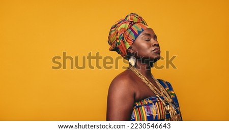 Ghanaian woman wearing traditional clothing against a yellow background. Mature black woman dressed in colourful Kente cloth and golden jewellery. Woman embracing her rich West African culture. Royalty-Free Stock Photo #2230546643