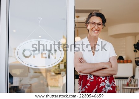 Happy cafe owner smiling at the camera while standing next to an open sign at the doorway of her coffee shop. Friendly small business owner welcoming customers to her newly opened restaurant.