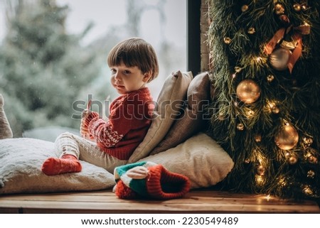 The kid sits at the window and waits for Santa Claus. Waiting for the Christmas holidays. Happy childhood. Waiting for firefighters.