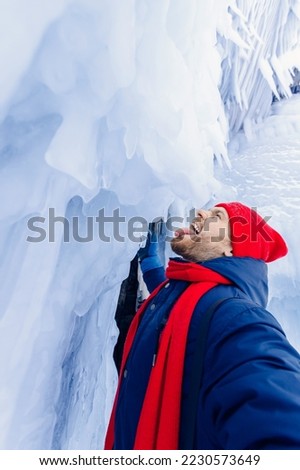 Adventure travel in winter, tourist man with red scarf makes selfie background ice grotto and cave, frozen icicles of Lake Baikal.