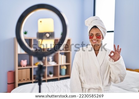 Hispanic woman with dark hair recording beauty tutorial with smartphone at home doing ok sign with fingers, smiling friendly gesturing excellent symbol 