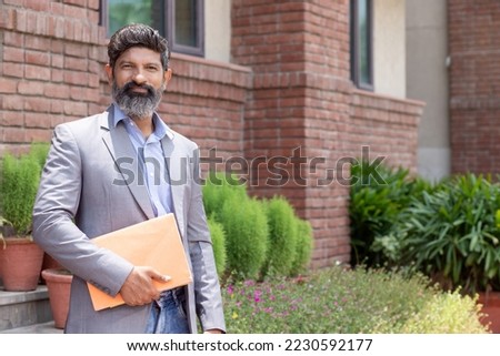 Portrait of happy indian mature professor of university or college or standing outdoor holding notebook in hand. Royalty-Free Stock Photo #2230592177