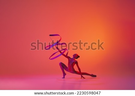Exercise with ribbon. Young flexible teen girl rhythmic gymnast in motion, action isolated over pink background in neon light. Sport, beauty, competition, flexibility, active lifestyle. Performance