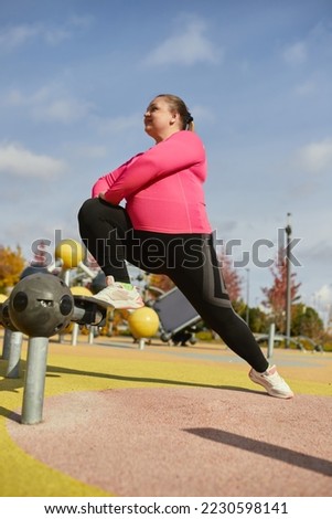Happy overweight young woman in colorful sportswear doing fitness exercises at street public sports ground, outdoors. Sunny autumn day. Concept of wellness, sport, health, mood, body positive