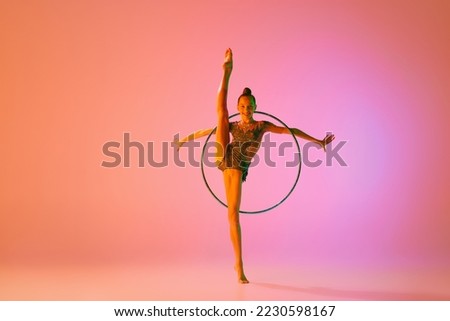 Studio shot of young charming girl, rhythmic gymnast training with sports equipment isolated over pink background in neon light filter. Dance, music, emotions, challenges, motivation and ad