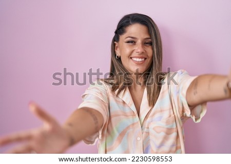 Blonde woman standing over pink background looking at the camera smiling with open arms for hug. cheerful expression embracing happiness. 