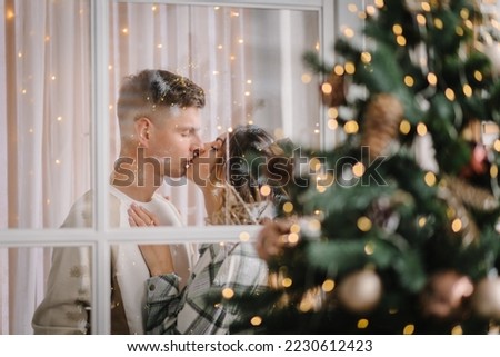 Wife hugs and kisses beloved husband near Christmas tree. Romantic couple in love spending eve together, woman and man enjoying winter vacations in cozy home interior. View through the window. Closeup