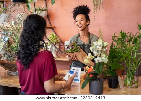 Young african florist holding bouquet of fresh flower while customer making digital payment with smartphone while scanning QR code. Black saleswoman giving a bunch of flower to customer at botany shop