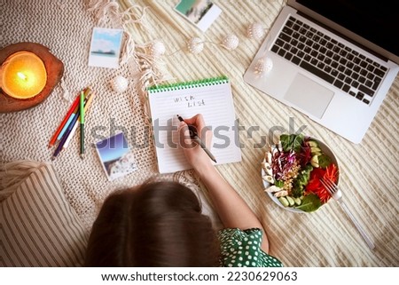 Top view of anonymous female writing wish list in notebook while lying on bed near laptop and power bowl