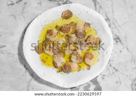 Scallop carpaccio with truffle mushroom slices in olive oil and herbs. Nearby is a glass of rosé sparkling wine and a bottle of sparkling wine. Food in a light ceramic plate on a marble countertop.