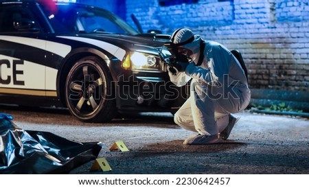 Forensics Specialist Taking Photos of Corpse in a Body Bag on a Crime Scene. Evening Shot of a Professional Police Team Officer in Coverall Suit Using Camera Documenting Clues and Traces