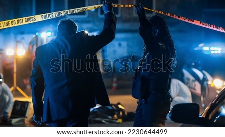 Team of Two Multiethnic Police Officers Working on Profiling a Killer on the Loose. Detectives on Duty at Night Solving Crime to Bring a Violent Murderer to Justice. Police Car and Paramedics on Site Royalty-Free Stock Photo #2230644499
