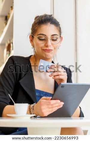 a dark-haired girl sitting at a table is holding her tablet and with a credit card is ready to make some purchases on the internet. The expression is one of uncertainty.