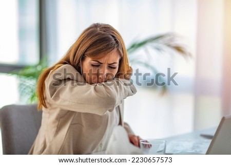 A beautiful business woman is sitting in the office, coughing in her elbow, correct behavior due to non-spreading viral infection. An unhealthy young woman feels bad at work, Covid 19 symptoms.