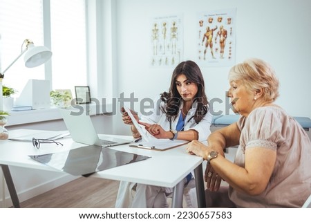 A female doctor sits at her desk and chats to an elderly female patient while looking at her test results. Patient Having Consultation With Female Doctor In Office