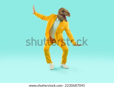 Funny, cheerful man wearing a dinosaur costume dancing and having fun. Happy guy in a bright yellow suit and a funny dinosaur mask dancing on a turquoise blue colour studio background. Party concept