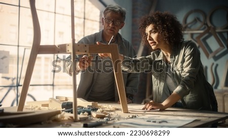 Portrait of Two Small Business Owners Working Together on Creating a New Wooden Dining Table Chair Design. Adult Man and Young Beautiful Female Looking at a Blueprint and Discussing the Work Process.