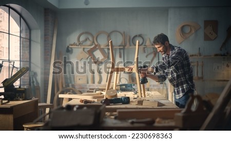 Young Carpenter Reading Blueprints and Starting to Assemble Parts of a Wooden Chair with a Rubber Hammer. Stylish Furniture Designer Working in a Studio in Loft Space with Tools on the Walls. Royalty-Free Stock Photo #2230698363
