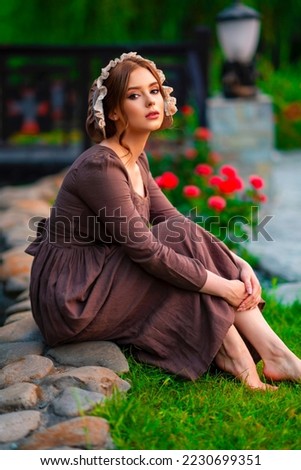 Beautiful red haired girl in brown long medieval dress and bonnet sitting on stone wall. Art work of romantic lady.Pretty tenderness model posing on nature. Royalty-Free Stock Photo #2230699351