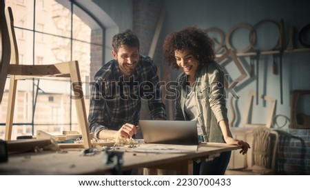 Happy Young Caucasian Man and Multiethnic Woman Using Laptop Computer in Designer Furniture Studio. Carpenters Have Fun, DIscussing and Talking About Upcoming Orders.